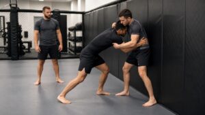 Two MMA trainees practicing a controlled wall-wrestling clinch while a coach observes
